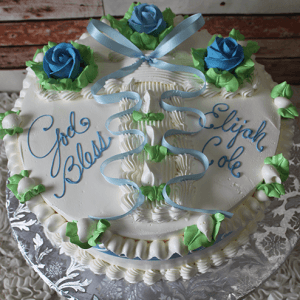 The RC-030 is a round white cake with blue and green icing roses, piped leaves, and a blue ribbon. “God Bless Elijah Cole” is written in blue script. The cake sits on a silver floral-patterned board.