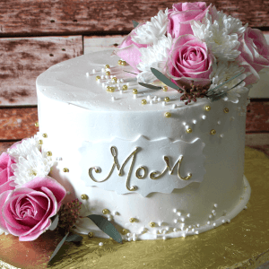 The MD-008 cake features white icing, decorated with pink roses, white chrysanthemums, gold and white pearls, and “Mom” written in gold icing. Presented on a gold board against a rustic wooden background.
