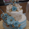 The TC-136 is a two-tier cake with blue and white flowers, gold leaves, a cross topper, and a gold cursive plaque, displayed on a patterned tablecloth.