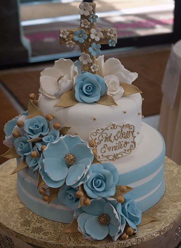 The TC-136 is a two-tier cake with blue and white flowers, gold leaves, a cross topper, and a gold cursive plaque, displayed on a patterned tablecloth.