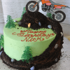 The SC-086 Dirt Bike is a green frosted cake featuring small trees, candy rocks, and a dirt bike topper riding up a chocolate dirt trail. "Happy Birthday Nick" is written in red icing.