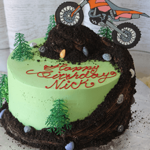 The SC-086 Dirt Bike is a green frosted cake featuring small trees, candy rocks, and a dirt bike topper riding up a chocolate dirt trail. "Happy Birthday Nick" is written in red icing.
