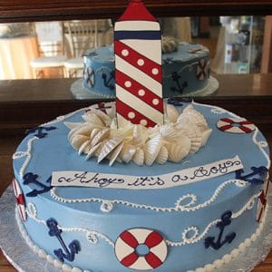 The BB-038 cake features a nautical blue design with white and red lifebuoys, navy anchors, rope icing, seashells, a tall lighthouse topper, and “Ahoy it’s a boy” written in script icing.