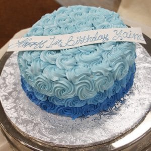 The CB-018B is a round cake with light and dark blue rosette icing, a white strip reading "Happy 1st Birthday Yaim" in blue cursive, served on a silver textured platter.