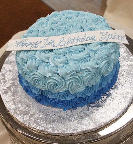 The CB-018B is a round cake with light and dark blue rosette icing, a white strip reading "Happy 1st Birthday Yaim" in blue cursive, served on a silver textured platter.