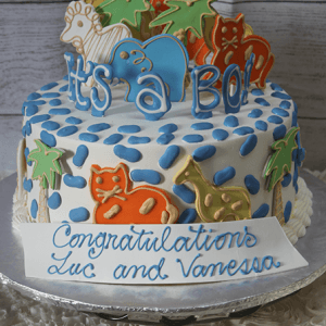 A round cake (BB-031) topped with animal cookies, blue and white icing, and “It’s a Boy!” A front sign says, “Congratulations Luc and Vanessa.”.