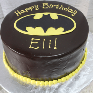The CB-006E is a round chocolate cake featuring a yellow Batman logo, “Happy Birthday Eli!” in yellow icing on top, and a border of yellow icing dots around the base.