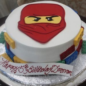 The CB-004A is a round birthday cake featuring colored Lego-like blocks on the sides and a red ninja face on top, with "Happy 5th Birthday Donovan" written in red script.