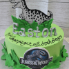 A green CB-014B Jurassic World cake features a cartoon dinosaur topper, green leaves, silver "Easton" letters, and "Happy Birthday" in black icing.