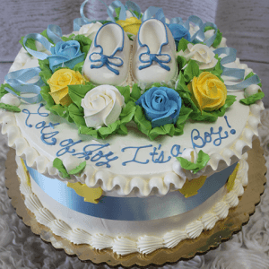 The BB-004 is a round cake adorned with blue, yellow, and white roses, topped with baby shoes, finished with a blue ribbon at the base, and features “Lots of Joy! It’s a Boy!” in blue icing.