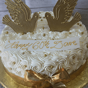 AN-021 is a round white cake with rose-like icing, gold pearls, and a gold ribbon bow. It features two gold dove toppers and a "Happy 60th Dave" message written in gold icing.
