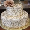 The WD-014 is a two-tier white cake with gold floral patterns, gold bead borders, and a large realistic tan fondant rose on top, presented on a gold patterned cake board.