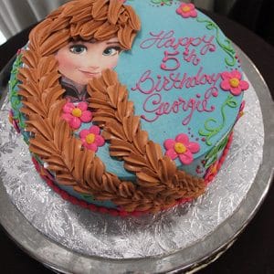 CB-008E is a round birthday cake with blue icing, decorated with a brown-haired character, pink flowers, and “Happy 5th Birthday Georgie” in pink icing. Presented on a silver platter.