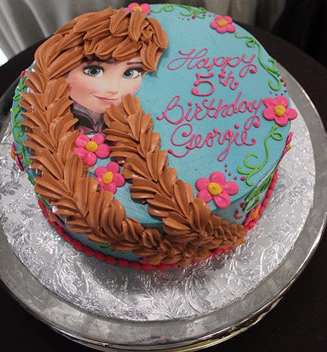 CB-008E is a round birthday cake with blue icing, decorated with a brown-haired character, pink flowers, and “Happy 5th Birthday Georgie” in pink icing. Presented on a silver platter.
