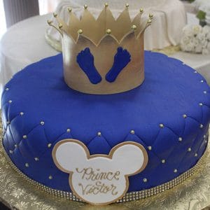 A round blue cake (BB-029) decorated with gold pearls, a gold crown with blue baby footprints, and a Mickey Mouse-shaped white plaque that reads “Prince Victor.”.