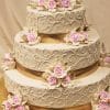 WC-057: Elegant three-tier wedding cake featuring intricate ivory swirls, gold ribbon accents, and clusters of light pink roses with cream leaves, presented on a gold cake board against a beige backdrop.