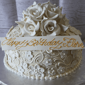 BR-009 is a white birthday cake featuring intricate floral icing, white sugar roses on top, and a white plaque with gold script that reads "Happy Birthday Flora.