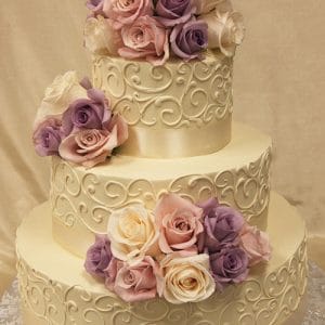 The WD-017 is a three-tiered ivory cake with ornate swirls, topped and adorned with clusters of pink, white, and purple roses, displayed on a patterned silver board against a beige background.