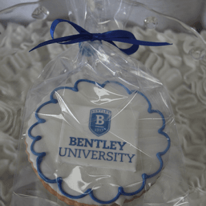 The GC-080M Bentley, a round cookie with the Bentley University logo and blue icing, is wrapped in clear plastic with a blue ribbon and displayed on ruffled white fabric.