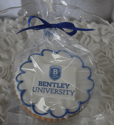 The GC-080M Bentley, a round cookie with the Bentley University logo and blue icing, is wrapped in clear plastic with a blue ribbon and displayed on ruffled white fabric.