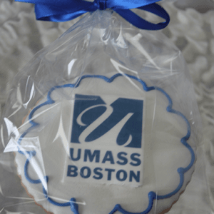 A GC-080B UMass Boston round cookie featuring the blue and white UMass Boston logo, wrapped in clear plastic and tied with a blue ribbon.