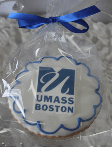A GC-080B UMass Boston round cookie featuring the blue and white UMass Boston logo, wrapped in clear plastic and tied with a blue ribbon.