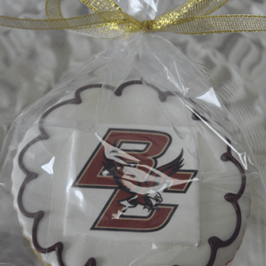 A round GC-080I Boston College cookie with white icing, a maroon border, and the Boston College logo with an eagle, wrapped in clear plastic and tied with a gold ribbon.