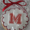 The GC-080J Milton High School cookie is wrapped in clear plastic with a red ribbon, featuring a large red M outlined in white and detailed with red icing around the edge.