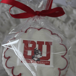 The GC-080L Boston University cookie is round with white icing, a red scalloped border, and features the BU logo and mascot. It's wrapped in clear plastic and tied with a red ribbon.