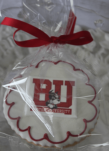 The GC-080L Boston University cookie is round with white icing, a red scalloped border, and features the BU logo and mascot. It's wrapped in clear plastic and tied with a red ribbon.