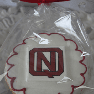 The GC-080D North Quincy High School is a round sugar cookie with white icing, a red border, and a red/black "N" in the center, wrapped in clear plastic with a red ribbon.