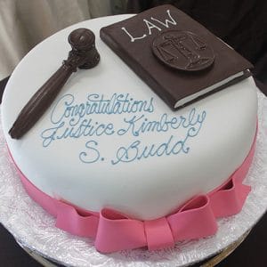 GC-052 is a round cake adorned with a pink ribbon, chocolate gavel, and chocolate law book, inscribed in blue icing with "Congratulations Justice Kimberly S. Budd.