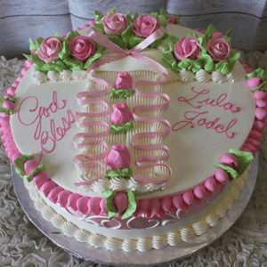RC-031 is a round white cake decorated with pink and green frosting roses, piped borders, and ribbons. “God Bless Lula Jodel” is written in pink icing on top, and the cake is presented on a silver board.
