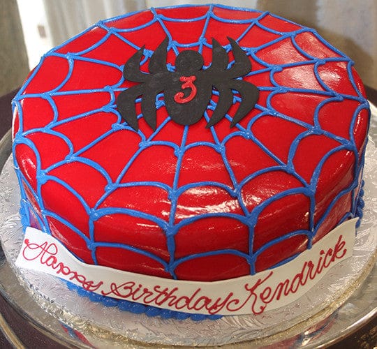 The CB-006A is a round cake with red frosting, blue web lines, and a black spider with a red number 3. A white banner at the base says "Happy Birthday Kendrick" in red cursive.