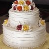 The WC-055 is a two-tiered cake with white frosting, wavy icing, and colorful fondant roses in yellow, orange, and maroon on top, plus matching flowers on the lower tier, all set on a gold base.