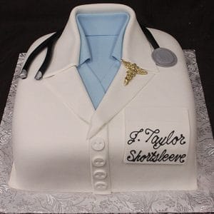 The BD-134 cake features a doctor’s white coat with blue shirt, stethoscope, caduceus pin, and a J. Taylor Shortsleeve name tag, all displayed on a patterned silver board.