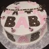 BB-020: Round cake featuring "BABY" in bold pink and brown letters, polka dots, fondant baby clothes on a clothesline, and small pink-brown fondant balls on a silver patterned board.