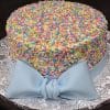 CB-029 is a round cake topped with colorful confetti sprinkles and finished with a large light blue fondant bow at the base, presented on a silver floral-patterned cake board.
