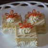 Three AP-045L Mimosa Squares—vanilla sponge cakes with cream filling, whipped cream, and orange decorative icing—are arranged on a white plate, each in a white paper liner.