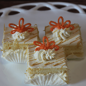 Three AP-045L Mimosa Squares—vanilla sponge cakes with cream filling, whipped cream, and orange decorative icing—are arranged on a white plate, each in a white paper liner.