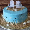 The BR-045 cake features blue icing, edible seashells, sand-like crumbs, two mini white Adirondack chairs on top, black birds and white clouds on the sides, and is presented on a gold tray.