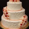 WC-059 is a three-tiered white wedding cake with intricate piping, peach icing borders, and clusters of pink, peach, and red roses. The bottom tier features the initials J.C.