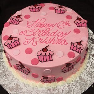 The CB-025C is a round pink birthday cake with pink and white polka dots, cupcake decorations, and "Happy 6th Birthday Krishna" in pink icing, presented on a silver-patterned board.