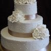 WC-052 is a three-tiered white wedding cake with quilted patterns, small pearl-like dots, champagne satin ribbons, and elegant white rose clusters on each layer.