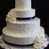 The WC-034 is a three-tiered white wedding cake featuring intricate scrollwork, dark blue ribbon bands on each layer, white sugar roses on the base and middle tier, and presented on a silver patterned board.