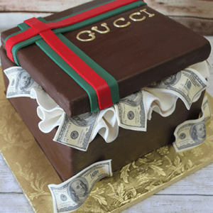 The BD-033C Designer Box RedGreen is a square cake styled as a Gucci gift box, featuring red and green ribbons, gold "GUCCI" lettering, ruffled white icing, edible $100 bills on the sides, and set on a gold board.