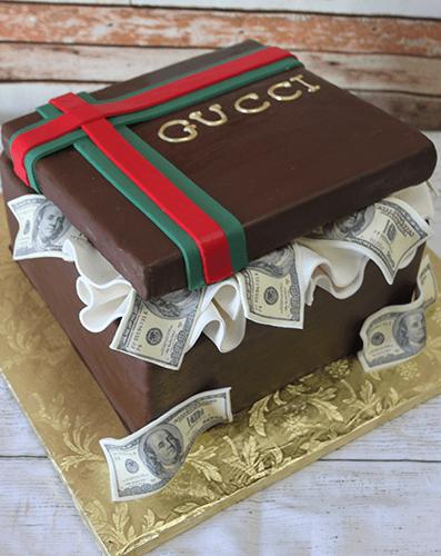 The BD-033C Designer Box RedGreen is a square cake styled as a Gucci gift box, featuring red and green ribbons, gold "GUCCI" lettering, ruffled white icing, edible $100 bills on the sides, and set on a gold board.
