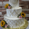 TC-023: A three-tier white cake with piped icing, sunflowers, pink roses, small white flowers, and greenery on a gold board, set against a rustic wooden background.