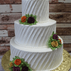 Product WC-024: A three-tiered white cake with textured diagonal lines, adorned with orange, purple, and yellow buttercream roses plus green leaves, presented on a gold floral-patterned board against a rustic wooden backdrop.
