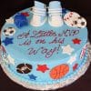 A round blue cake (BB-025) features fondant sports balls, stars, a blue cap, baby sneakers on top, and red icing that says “A Little MVP is on his Way!”.